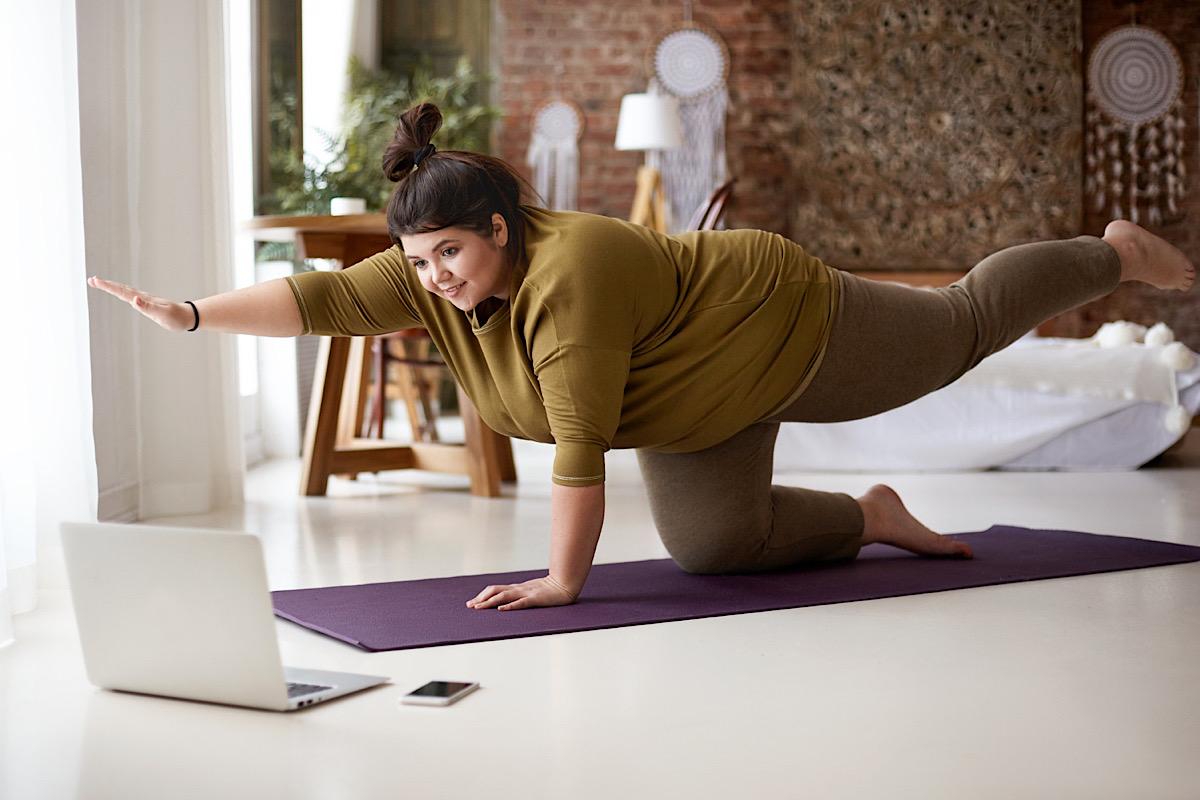 Füllige Frau ist auf der Yogamatte im Vierfussstand, ein Arm und ein Bein ausgestreckt. Sie schaut auf ihren Laptop.
