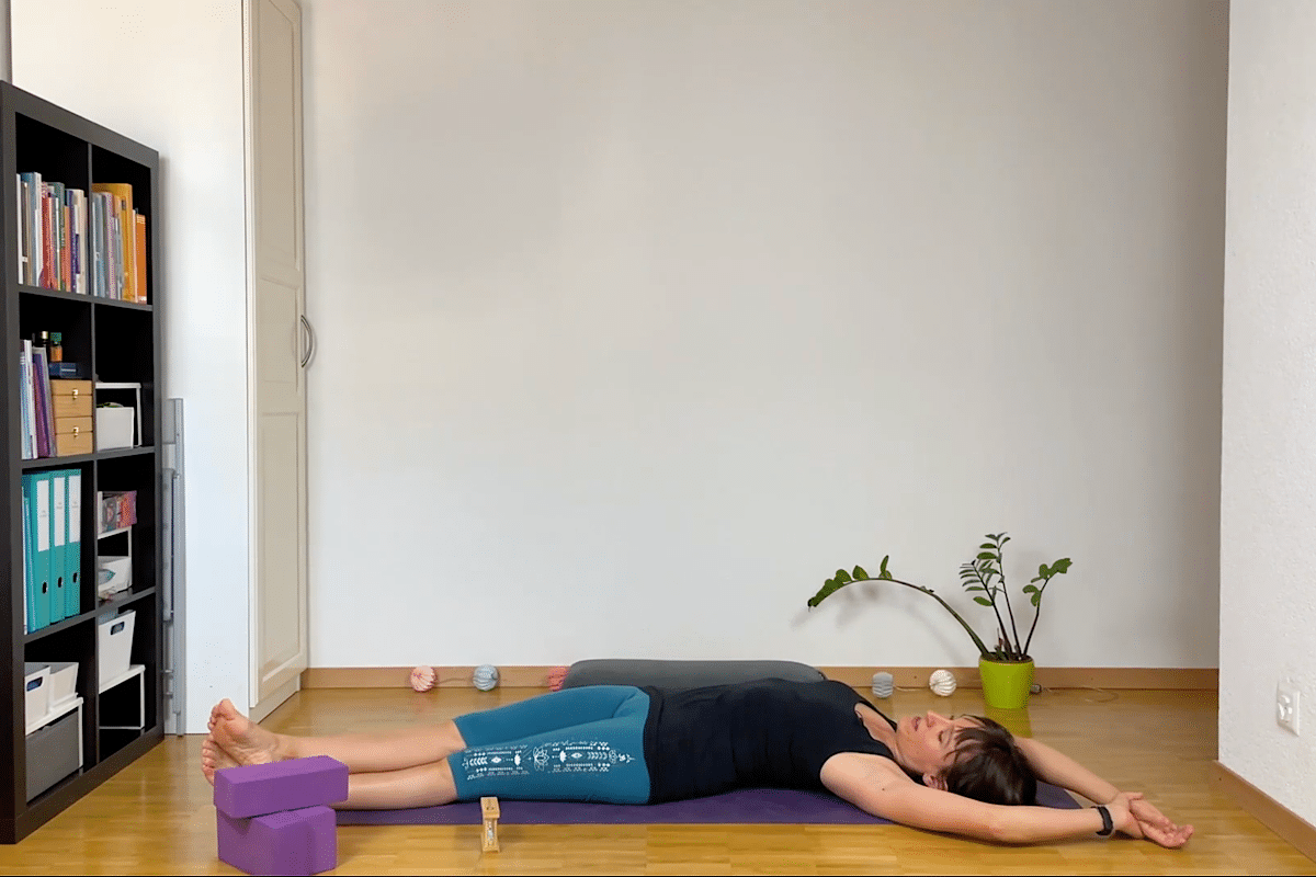 Katharina Balande demonstriert den liegenden Halbmond: auf dem Rücken liegen, Beine und Arme gestreckt, eine C-Form machen und so eine Seite dehnen.