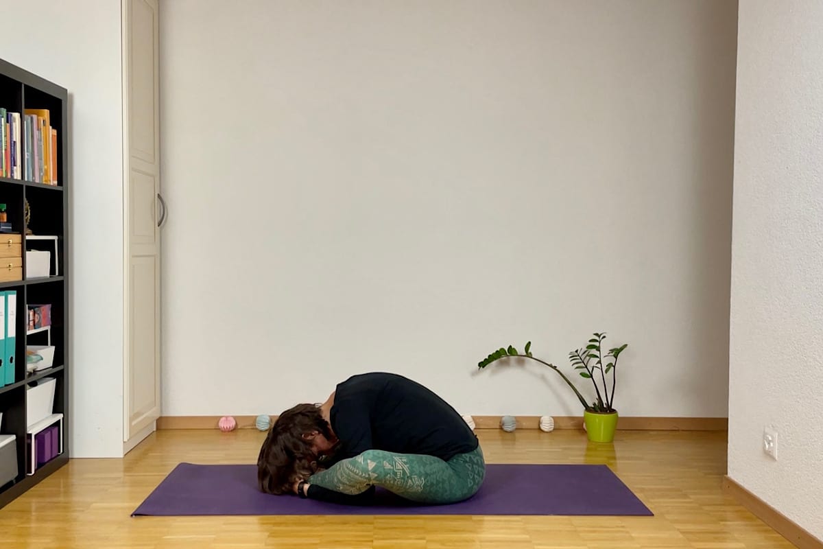 Katharina Balande in der Stellung "Schildkröte". Sie sitzt auf der Yogamatte. Die Beine sind in Rautenform vor dem Körper. Füsse berühren einander. Die Arme sind unter den Beinen und die Hände fassen die Füsse. Oberkörper und Kopf hängen locker.