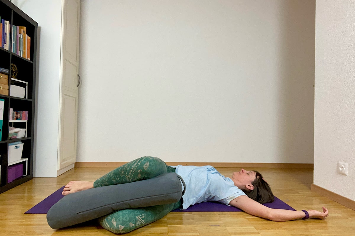 Katharina Balande in der Yin Yoga Stellung Korkenzieher. Rückenlage, Beine gebeugt, Knie aufeinander, fallen zu einer Seite, dazwischen ein Kissen, Arme über den Kopf.