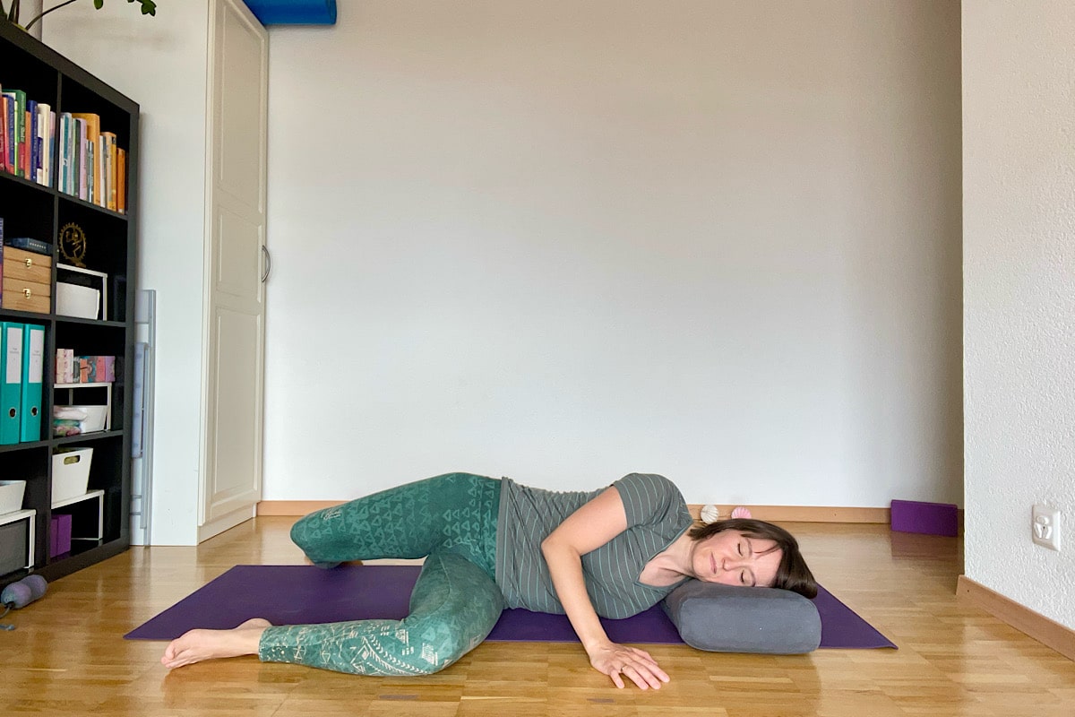 Katharina Balande zeigt Yin Yoga Stellung offene Flügel. Seitlage, ein Arm zur Seite weggesteckt. Beide Knie im 90° angewinkelt, unteres Bein zusätzlich im 90° angewinkelt.