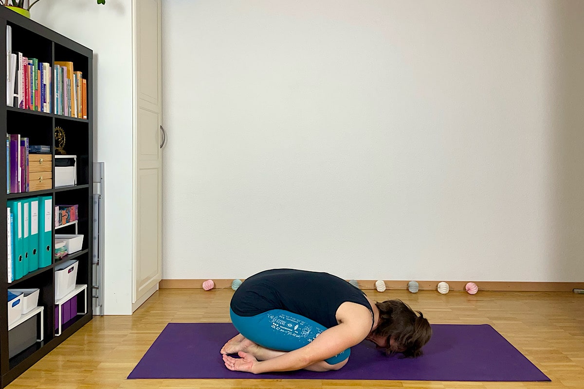 Eine Person praktiziert Yoga im Sommer auf einer lila Matte in einem Raum mit Holzboden. Sie führt die Stellung des Kindes aus, mit gesenktem Kopf und Armen an den Seiten. Im Hintergrund steht ein Bücherregal mit Büchern und Ordnern, das den ruhigen Übungsraum vervollständigt.