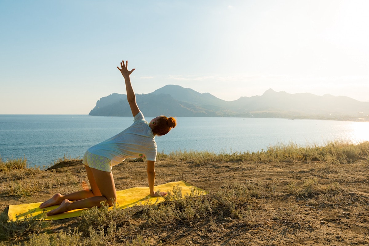 Eine Person praktiziert im Sommer Yoga auf einer gelben Matte im Freien auf einer Rasenfläche mit Blick auf ein ruhiges Gewässer und Berge. Der Himmel ist klar und die Sonne scheint hell.