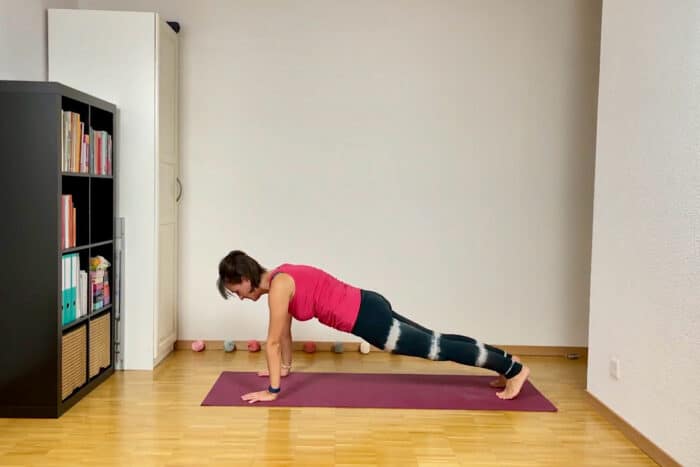 Eine Person in einem roten Tanktop und schwarzen Leggings hält auf einer lila Yogamatte in einem hellen Raum eine Plank-Position, die Teil des Sonnengruss im Yoga ist. Links steht ein schwarzes Bücherregal voller Bücher und Kleinkram.