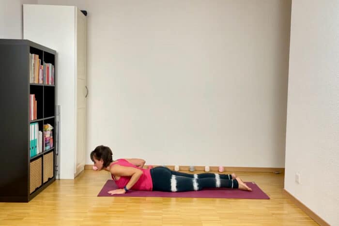 Eine Person in einem rosa Tanktop und schwarzen Batik-Leggings führt anmutig die Kobra-Pose aus. Sie ist Teil des Sonnengruss im Yoga. Links trägt ein schwarzes Bücherregal voller Bücher zur heiteren Atmosphäre bei.