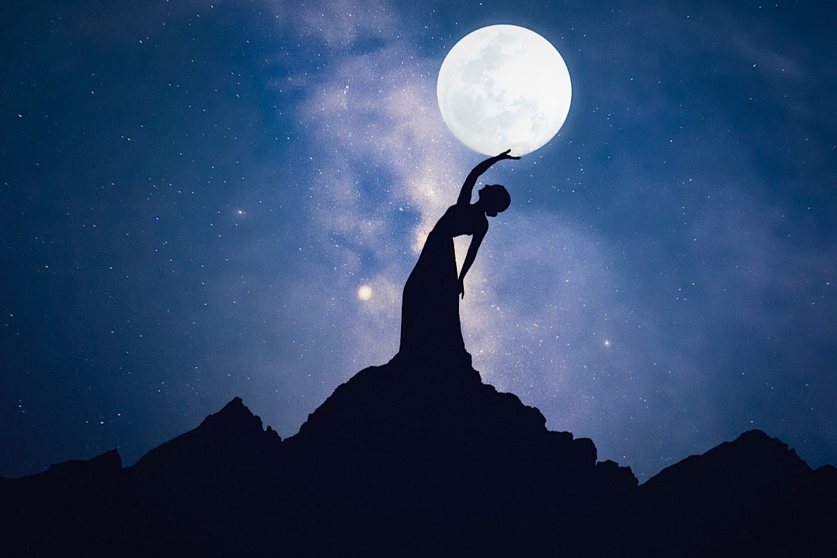 Die Silhouette einer Person auf einem Berggipfel streckt sich nach einem großen, hellen Vollmond vor einem sternenklaren Nachthimmel und ruft die heitere Ausgeglichenheit hervor, die man beim Yoga in Hitze finden kann.