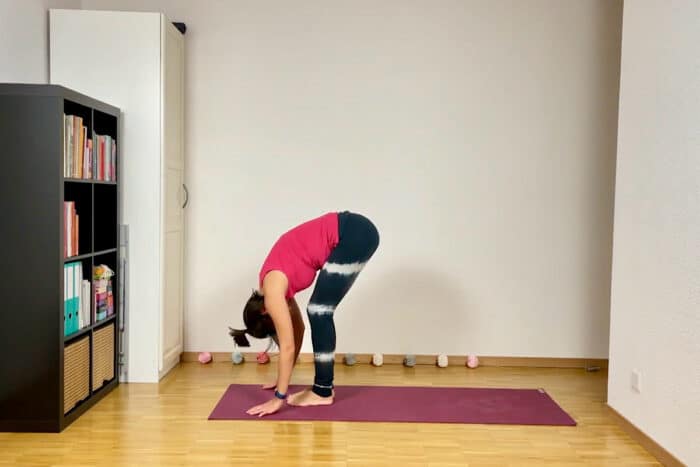 Eine Person in einem roten Tanktop und schwarzen Leggings praktiziert in einem hellen Raum den Sonnengruß im Yoga. Sie befindet sich in einer Vorwärtsbeuge auf einer roten Yogamatte. Im Hintergrund ist ein schwarzes Bücherregal voller Bücher und ein paar bunte Yogablöcke auf dem Boden.