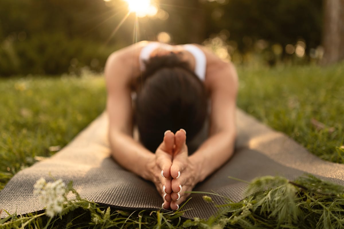 Eine Person in einem weißen Tanktop führt die Kinderpose, ein typisches Yin-Yoga-Element, auf einer Matte im Freien aus. Die untergehende Sonne verbreitet warmes Licht und schafft eine friedliche Atmosphäre, während die wohltuende Tiefenwirkung genutzt wird. Gras und kleine Blumen umgeben die Matte.