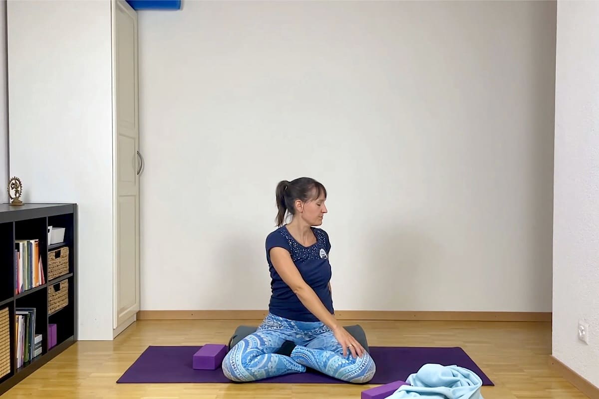 Eine Frau sitzt im Schneidersitz auf einer Yogamatte und dreht ihren Oberkörper nach links, wobei sie ihre rechte Hand auf ihr linkes Knie legt.