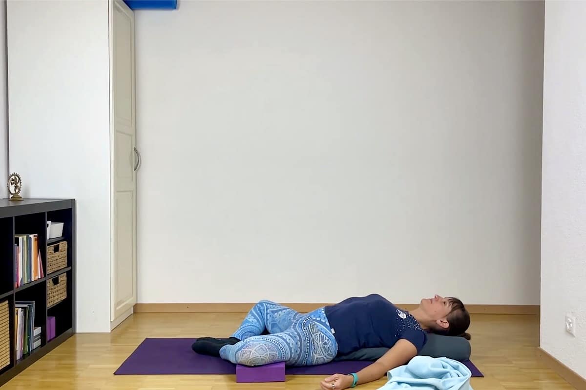 Eine Person liegt auf einer Yogamatte in der zurückgelehnten Schmetterlingspose mit einem Kissen unter dem Rücken, einem Block unter dem Knie.
